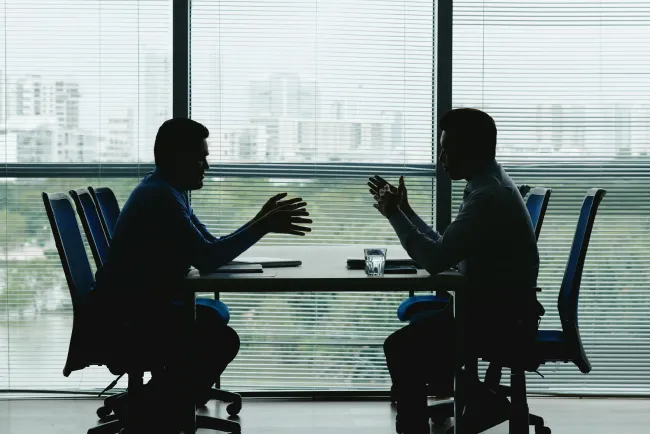 two-human-outlines-against-shuttered-office-window-sitting-opposite-each-other-negotiating