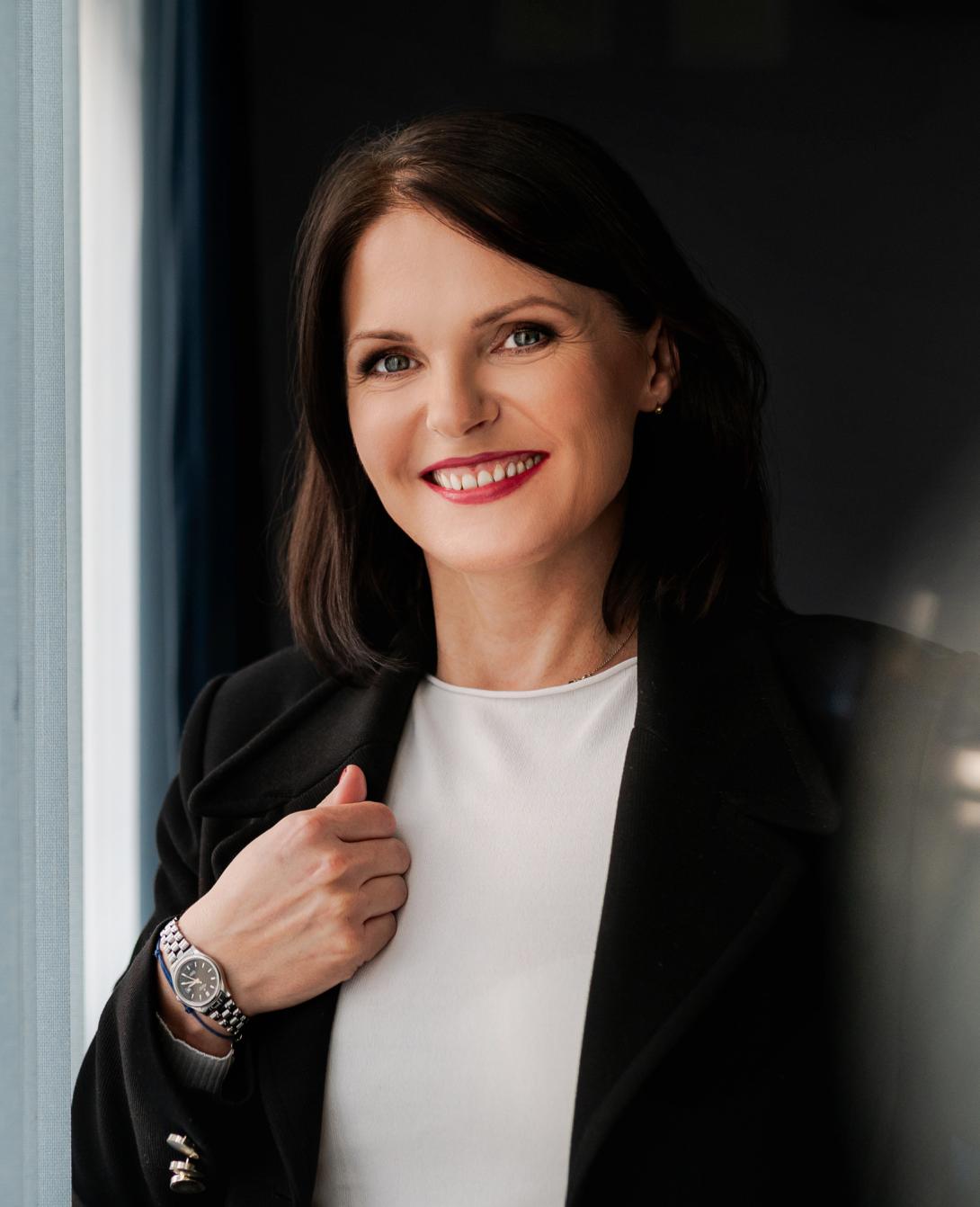 Uśmiechnięta kobieta o ciemnych włosach, ubrana w czarną marynarkę i białą bluzkę, pozuje z dłonią przy klapie żakietu, na której widoczny jest zegarek.