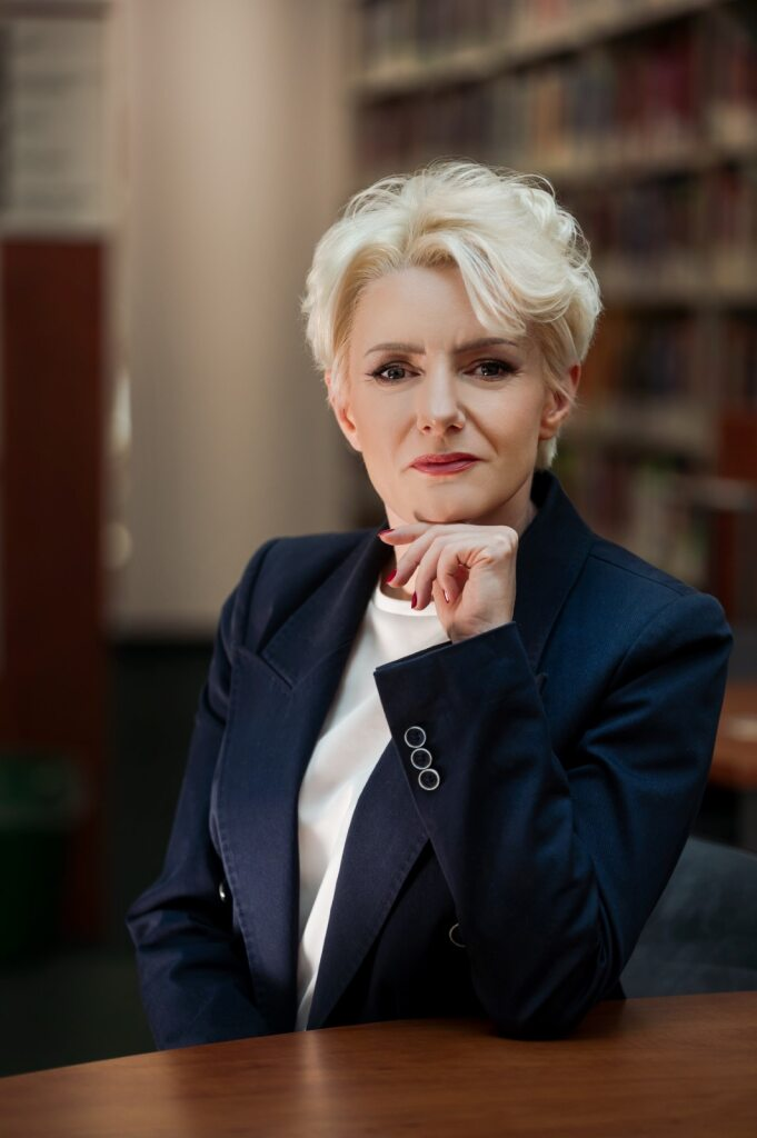 Zdjęcie kobiety w ciemnej marynarce i białej bluzce, siedzącej przy stole na tle regałów z książkami.