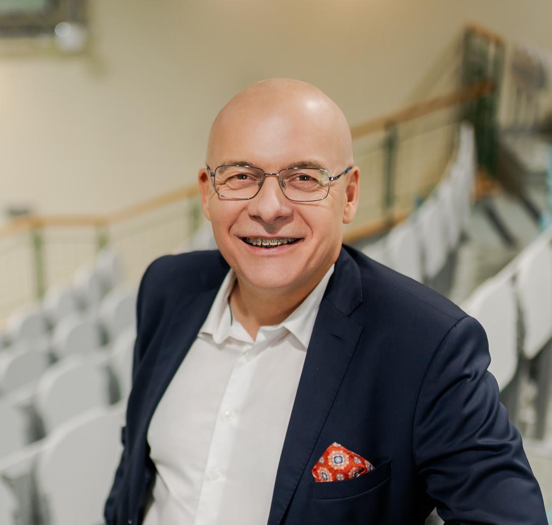 Uśmiechnięty, łysy mężczyzna w okularach i granatowej marynarce z wzorzystą poszetką pozuje na tle rzędów jasnych foteli w auli.