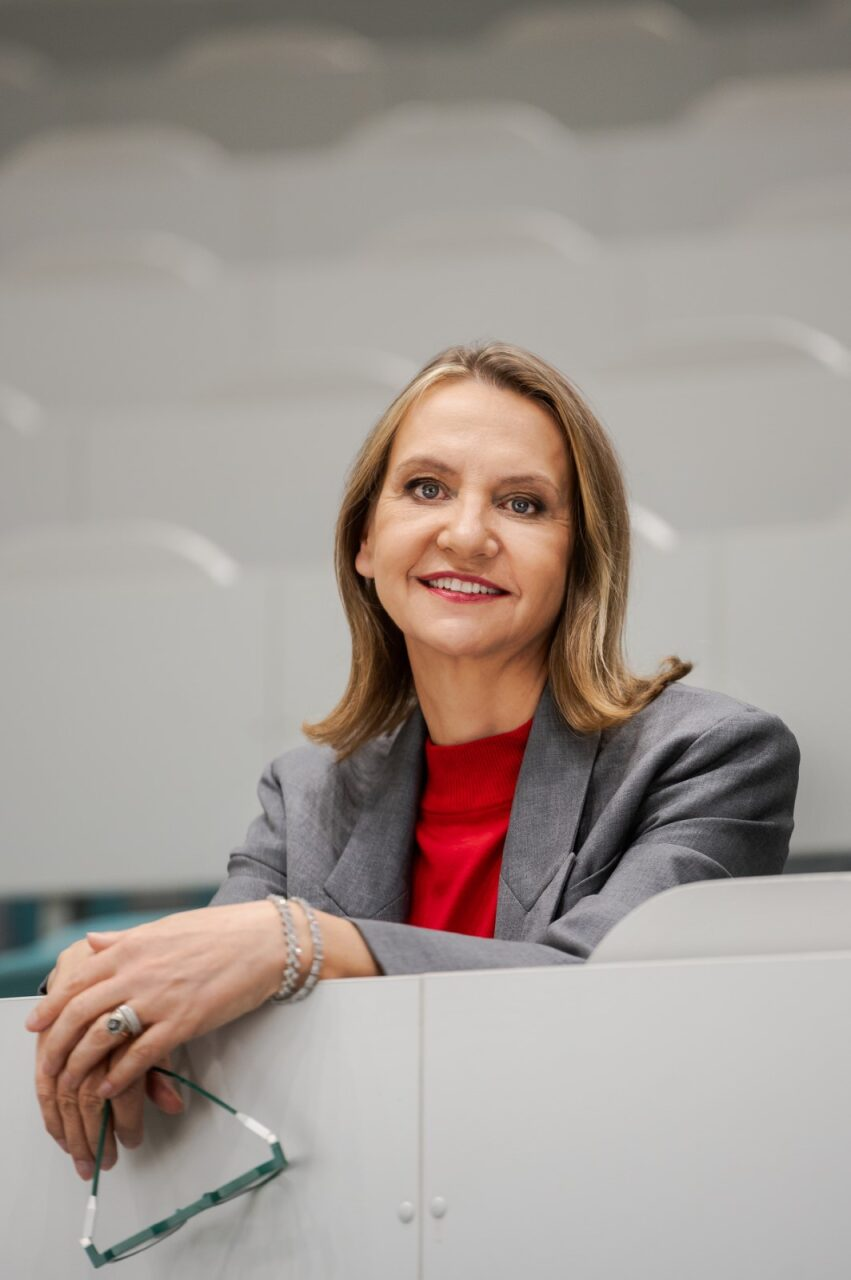 Uśmiechnięta kobieta w szarym żakiecie i czerwonym golfie opiera się o biały pulpit, trzymając w dłoni okulary na tle rzędów jasnych foteli w auli.